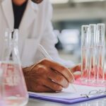 A scientist in a lab coat writes notes on a clipboard beside test tubes filled with clear liquid, with laboratory glassware visible on the table.
