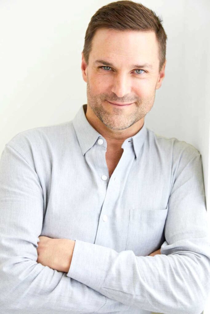 A man with short brown hair and light stubble smiles at the camera with his arms crossed, wearing a light gray button-up shirt against a white background.