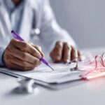 A person in a white coat writes on a document with a purple pen at a desk. A pair of eyeglasses and a stethoscope are placed on the table nearby. The scene suggests a medical or professional setting.