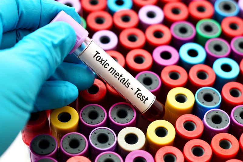 A gloved hand holds a blood sample tube labeled "Toxic metals Test" above a collection of colorful, capped test tubes.