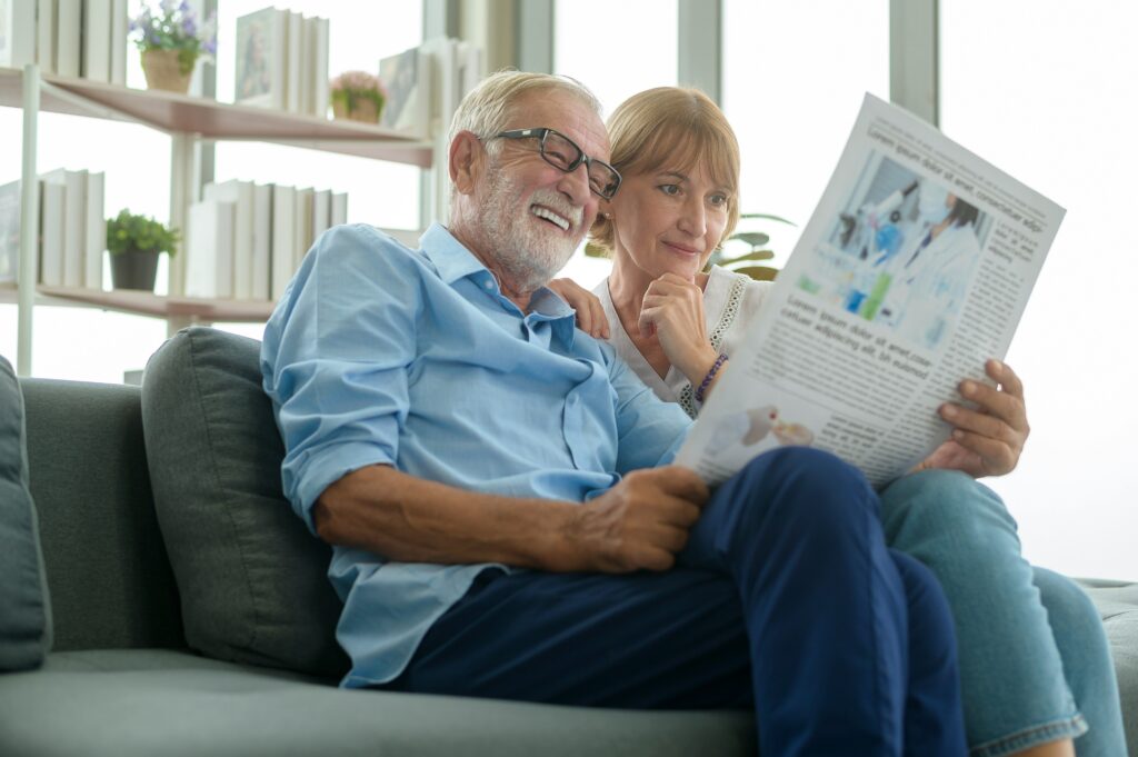 An older man and woman sit together on a couch, smiling as they read a newspaper. Books and potted plants are visible on shelves in the background, and natural light brightens the cozy living room.