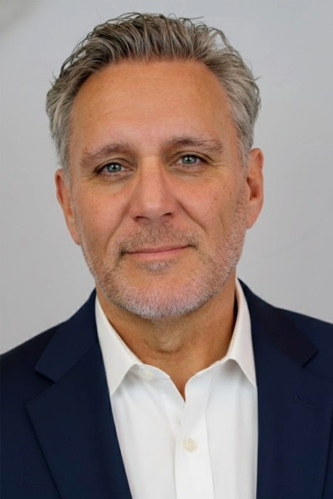 A middle aged man with short gray hair and a trimmed beard is wearing a navy blue suit jacket and a white collared shirt, smiling slightly against a plain light background.