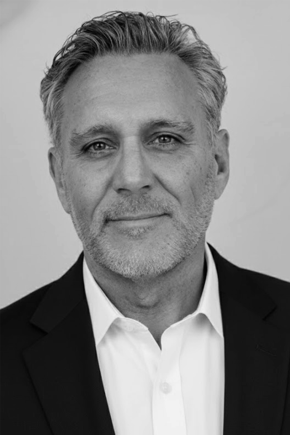 A middle aged man with gray hair and a neatly trimmed beard, wearing a dark suit jacket and white shirt, looks into the camera with a slight smile. The photo is in black and white.