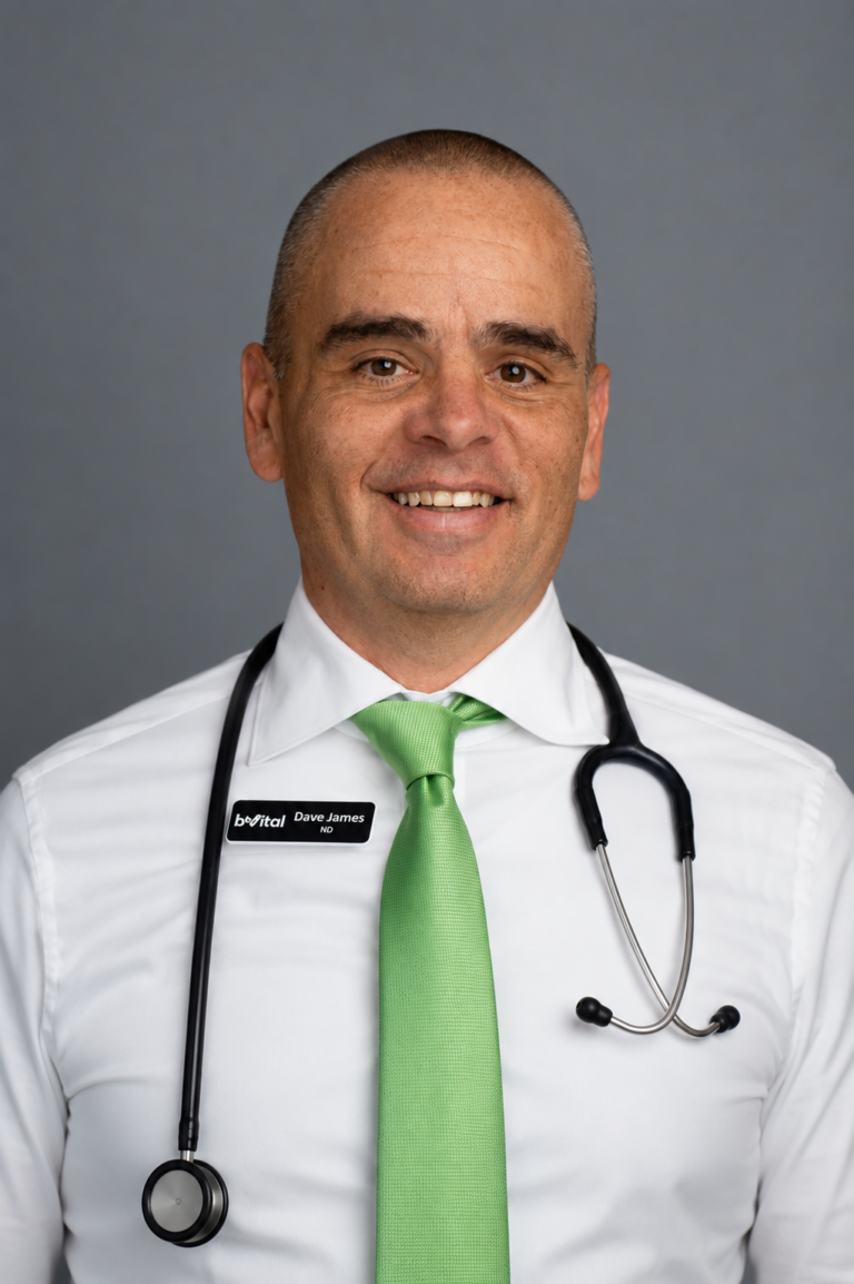 A smiling man wearing a white shirt, light green tie, and stethoscope, with a name tag that reads "Dr. James." He stands against a plain gray background.