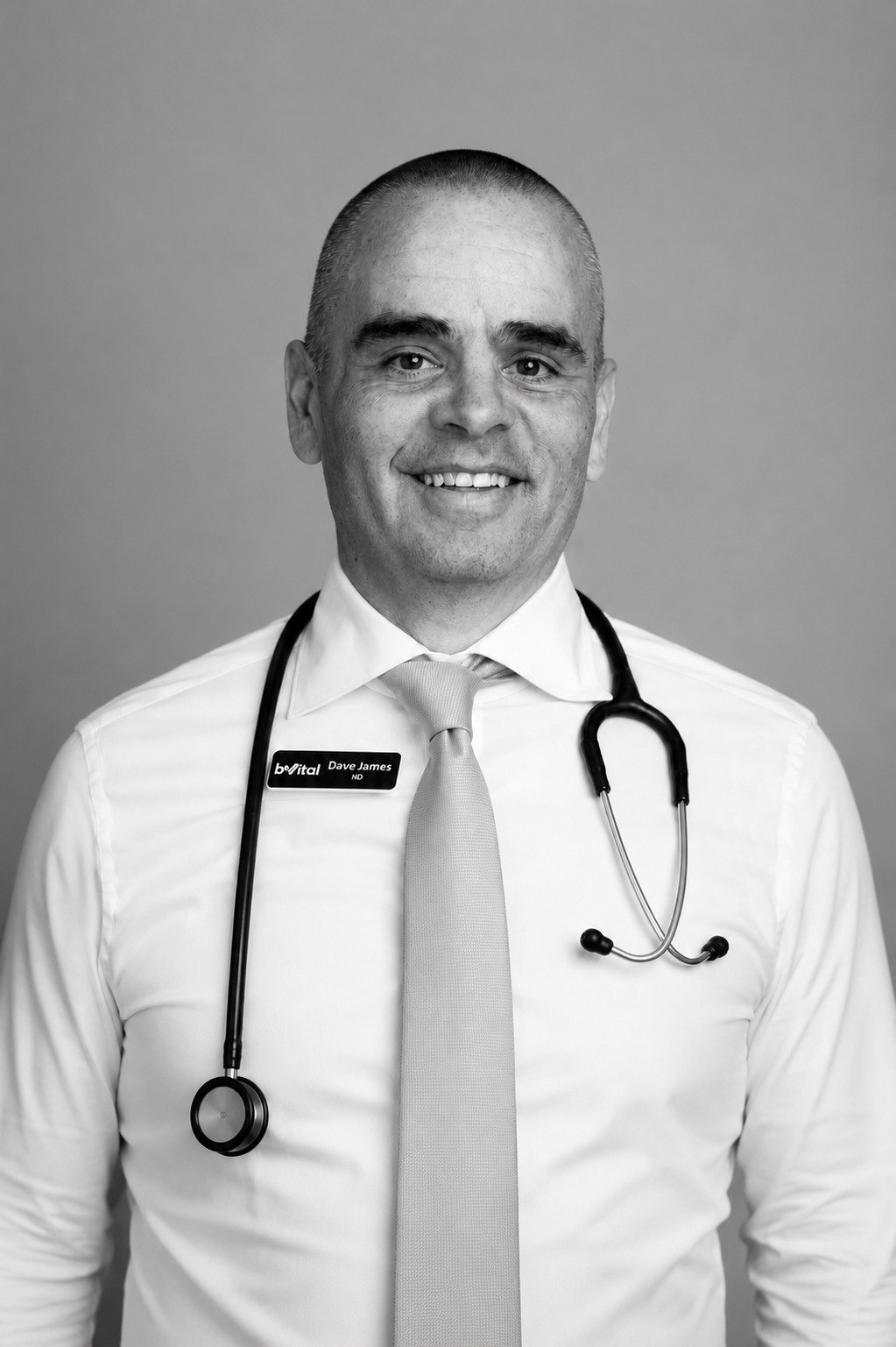 A smiling man in a shirt and tie wears a stethoscope around his neck and a name badge. The photo is black and white with a plain background.