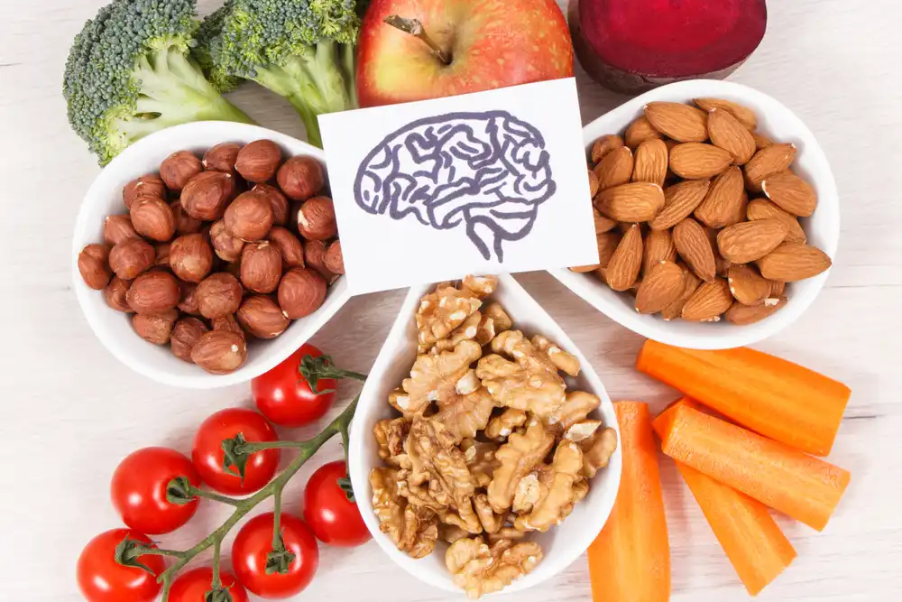 Bowls of hazelnuts, almonds, and walnuts arranged around broccoli, an apple, beet, cherry tomatoes, and carrot sticks, with a card showing a brain illustration in the center.