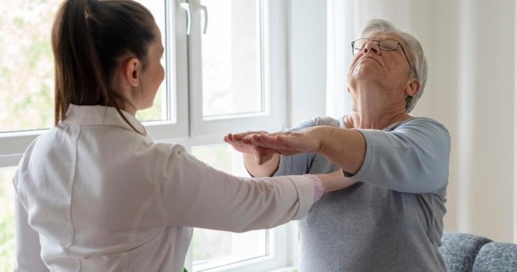 A younger woman assists an older woman with stretching exercises indoors. The older woman has her arms extended forward while the assistant supports her, both standing near a large bright window.