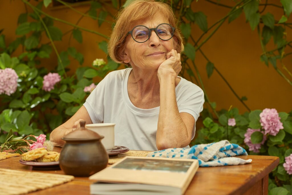 An elderly woman with glasses sits at a wooden table outdoors, smiling with her chin resting on her hand. There are a teapot, cup, plate of cookies, towel, and book on the table. Pink flowers and green plants are in the background.