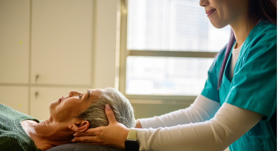 A healthcare worker in scrubs gently supports the head and neck of an older adult lying on a bed, in a brightly lit room with large windows in the background.