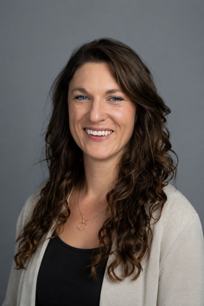 A woman with long curly brown hair, wearing a beige blazer over a black top, smiles at the camera in front of a plain gray background.