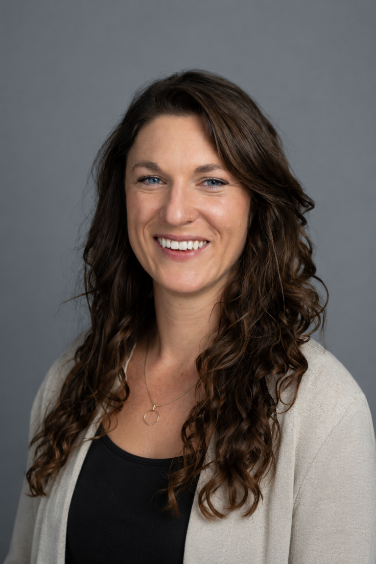 A woman with long curly brown hair, wearing a beige blazer over a black top, smiles at the camera in front of a plain gray background.