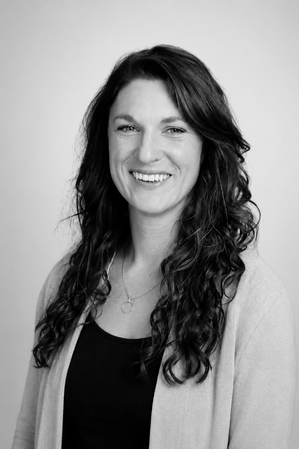 A woman with long, wavy hair smiles at the camera. She is wearing a light cardigan over a dark top and a simple necklace. The photo is black and white with a plain background.